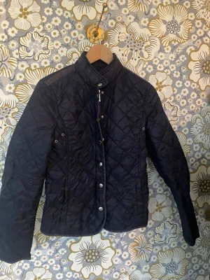 Mörkblå quiltad jacka Tommy Hilfiger - Snygg mörkblå quiltad jacka från Tommy Hilfiger i storlek M men sitter bra på även folk som har S Jackan har ståkrage, tryckknappar framtill och coola detaljer med metallknappar. Den är figursydd och har lång ärm, perfekt för dig som gillar klassisk stil med en modern twist.