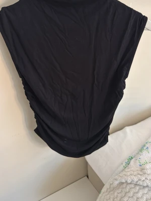 Svart ärmlös topp Gina Tricot  - Säljer en svart, ärmlös topp från Gina Tricot i storlek S. Toppen har en enkel och stilren design med lätt drapering i sidorna och mjukt, stretchigt material. Den har en liten fläck på sig (bild 3)!