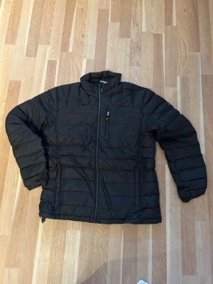 Svart pufferjacka från McKinley - Säljer en svart pufferjacka från McKinley med hög krage och dragkedja framtill. Jackan har en bröstficka med dragkedja och två sidofickor. Perfekt för kalla dagar och enkel att matcha med allt. Klassisk quiltad look och diskret logga på ärmen.