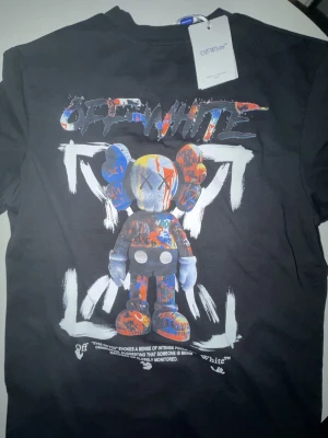  Off-White t-shirt  - Svart t-shirt från Off-White med stor färgglad grafisk print på ryggen och mindre tryck på bröstet. Motivet är en streetart-inspirerad figur i blått, rött, orange och vitt. Klassisk rund hals och korta ärmar. Materialet är mjuk bomull.