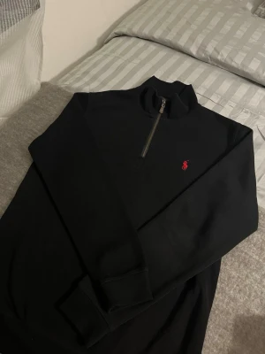 Svart half zip tröja från Polo Ralph Lauren - Svart långärmad tröja från Polo Ralph Lauren med röd broderad logga på bröstet. Tröjan har en half zip-dragkedja och hög krage. Tillverkad i mjuk bomull, perfekt för lager-på-lager under höst och vinter.