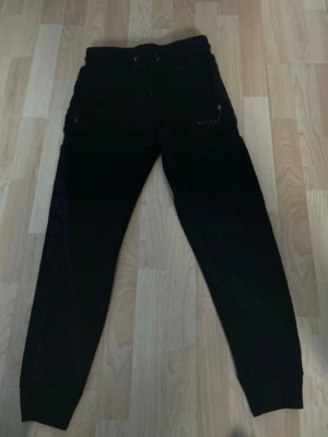 Svarta mjukisbyxor från McKenzie - Svarta mjukisbyxor från McKenzie med dragsko i midjan och dragkedjor på fickorna. Byxorna har en slim passform och är gjorda i mjukt material som är perfekt för chill eller träning. Snygg logga på ena benet och ribbade muddar vid bensluten. Storlek M