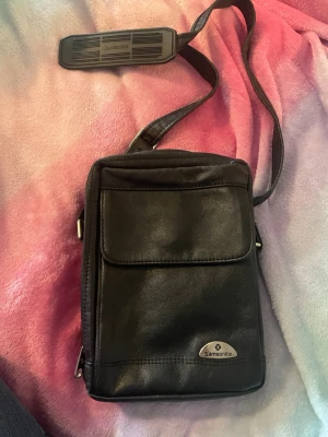 Svart axelväska i skinn från Samsonite - Snygg svart axelväska från Samsonite i äkta skinn. Väskan har justerbar axelrem, flera fack med dragkedja och smarta kortfack på insidan. Perfekt för dig som vill ha koll på grejerna och gillar stilren design.