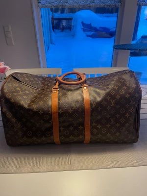 Louis Vuitton Keepall Monogram 60 resväska - Säljer en klassisk Louis Vuitton Keepall 60 i monogram canvas med bruna skinnhandtag och detaljer. Väskan har det ikoniska LV-mönstret i brunt och beige, dragkedja upptill och förstärkta hörn. Perfekt storlek för weekendresor och riktigt snygg att bära.