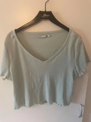 Ljusgrön ribbad croppad topp 157 - Superfin ljusgrön croppad topp från 157 med v-ringning och korta ärmar. Toppen är ribbad och har vågig kant nertill och vid ärmslut, vilket ger en extra snygg detalj. Perfekt för varmare dagar och enkel att matcha med jeans eller kjol.
