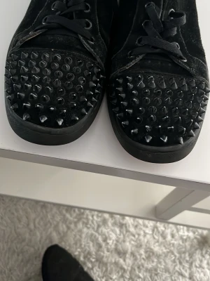 Svarta sneakers med nitar och röd sula - Unika svarta sneakers i mocka från Christian Louboutin med coola svarta nitar på tån och klassisk röd sula. Skorna har snörning och rund tå, samt en lyxig känsla med skinnfoder. Perfekta för dig som vill sticka ut med edgy detaljer.