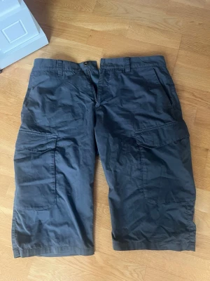 Svarta cargoshorts Peak Performance - Säljer ett par svarta cargoshorts från Peak Performance i storlek 34. Shortsen har stora sidofickor och är tillverkade i en mix av bomull och polyamid för en skön och slitstark känsla. Perfekta för dig som gillar praktiska och stilrena shorts.