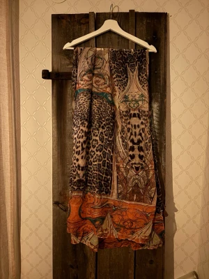 Leopardmönstrad sjal från By Jolima - Säljer en tunn och flowy sjal från By Jolima med leopardmönster och coola detaljer i orange, beige och svart. Västen har ett bohemiskt print med inslag av turkos och brunt, perfekt för att lyfta vilken outfit som helst.