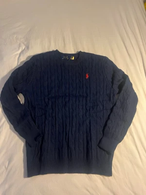 Mörkblå kabelstickad tröja Polo Ralph Lauren - Kabelstickad mörkblå tröja från Polo Ralph Lauren med klassisk röd broderad logga på bröstet. Rund halsringning och långa ärmar. Perfekt till höst och vinter för en stilren look. Sitter som en L