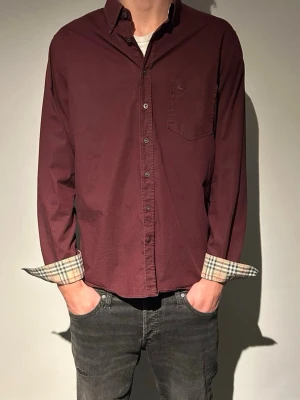 Vinröd burberry skjorta - Säljer en vinröd skjorta från Burberry med klassiska rutiga muddar och bröstficka. Skjortan har knappar hela vägen och en avslappnad passform. Perfekt för dig som vill ha en stilren men ändå unik look.