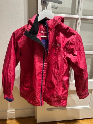 Röd vindjacka från Everest - Röd vindjacka från Everest med huva och blått meshfoder. Jackan har dragkedja framtill, justerbara ärmslut med kardborre och reflekterande detalj på huvan. Perfekt för blåsiga dagar och enkel att packa ner i väskan.