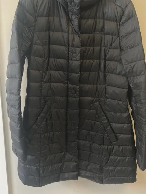 Svart lång pufferjacka från Everest - Säljer en svart lång pufferjacka från Everest med quiltad design och praktiska fickor. Jackan har dragkedja och knappar framtill, samt resår i midjan för snygg passform. Perfekt för kalla dagar och riktigt skön att bära.