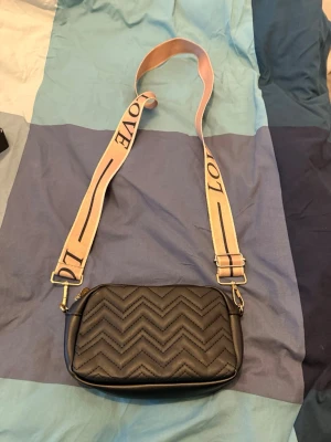 Svart quiltad axelväska med LOVE-band - Säljer en svart axelväska i quiltad design med zigzag-mönster. Väskan har ett brett, beige axelband med texten LOVE i svart. Materialet är syntet och väskan stängs med dragkedja. Perfekt storlek för det viktigaste och ger en cool vibe till din outfit.