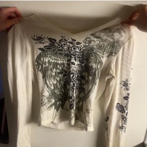 Vit långärmad topp - magtröja  - Säljer en vit ribbad långärmad topp med svart mönster framtill och på ärmarna. Toppen har en v-ringad hals och är croppad i modellen. 