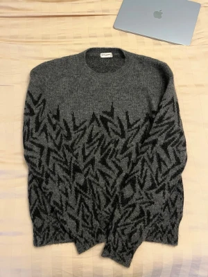 Grå stickad Saint Laurent tröja L - Säljer en grå stickad tröja från Saint Laurent i storlek L. Tröjan har ett svart grafiskt mönster över hela framsidan och ärmarna. Rund halsringning och ribbade muddar. Tillverkad i Italien, perfekt för dig som gillar exklusiva och snygga plagg. Brand lable är lite av men kan fixas enkelt