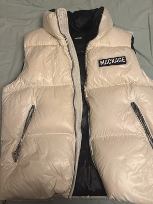 Vit dunväst från Mackage - Säljer en riktigt snygg vit dunväst från Mackage med hög krage och två stora dragkedjefickor. Västen har en tydlig logga på bröstet och quiltad design som ger en puffig look. Perfekt att slänga över en hoodie för extra värme.