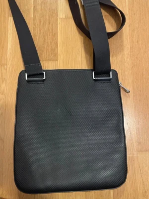 Svart axelväska från Lacoste - Säljer en stilren svart axelväska från Lacoste med det klassiska krokodilloggan framtill. Väskan har justerbar axelrem, diskret struktur i materialet och dragkedja upptill. Perfekt för dig som vill ha en clean och praktisk väska till vardagen.