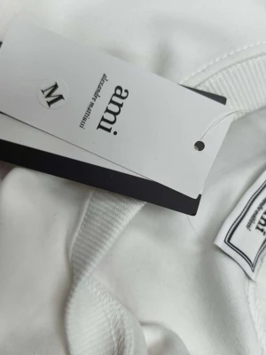 Vit sweatshirt från Ami med rött hjärta - Säljer en clean vit sweatshirt från Ami Alexandre Mattiussi i storlek M. Tröjan har ett stort rött hjärta med A-bokstav broderat på bröstet och ribbade muddar vid hals, ärmslut och nederkant. Perfekt för dig som gillar stilrena plagg med en twist.