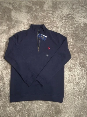 Marinblå half zip från Polo Ralph Lauren - Säljer en marinblå långärmad tröja från Polo Ralph Lauren med klassisk röd logga på bröstet. Tröjan har en snygg half zip-dragkedja och står för en clean och tidlös stil. Perfekt till jeans eller chinos för en chill look.