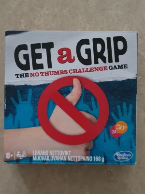 Get a Grip - The No Thumbs Challenge Game - Get a Grip är ett roligt sällskapsspel där du ska klara utmaningar utan att använda tummarna. Spelet innehåller tumhandskar med rött och vitt mönster, modellera, kort och tillbehör. Perfekt för att utmana vänner och familj på kreativa och knasiga uppgifter.