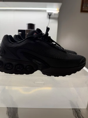 Nike Air Max DN - Säljer ett par helsvarta Nike Air Max sneakers med mesh-överdel och coola detaljer på sulan. Skorna har snörning, rund tå och en chunky sula med synliga luftbubblor. Perfekta för dig som gillar sportig och modern stil.