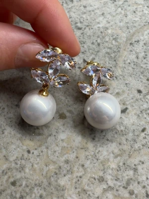 CC örhängen  - Säljer ett par eleganta örhängen med stora vita pärlor och gnistrande kristaller i bladform. Örhängena har guldfärgade detaljer som ger en lyxig känsla. Perfekta för dig som vill sticka ut med något klassiskt men ändå glam.