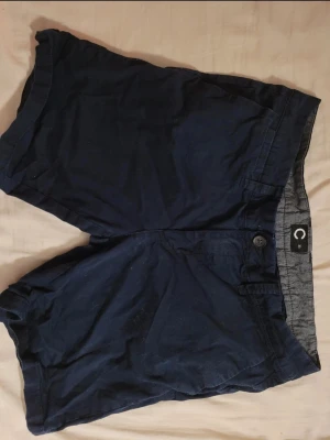 Mörkblå shorts med knapp - Säljer ett par mörkblå shorts i klassisk modell med knapp och gylf framtill. Shortsen har en enkel design och är tillverkade i ett mjukt bomullstyg som känns skönt mot huden. Perfekta för varma dagar och enkla att matcha med olika toppar.