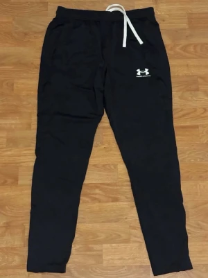 Svarta mjukisbyxor från Under Armour - Svarta mjukisbyxor från Under Armour med vit dragsko i midjan och logga på låret. Byxorna har en avslappnad passform och är perfekta för chill eller träning. Tillverkade i mjukt material för extra komfort.