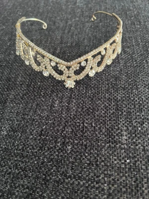 Glittrig tiara i guld och silver - Säljer en glittrig tiara i guld med massor av gnistrande stenar och detaljerad design. Perfekt accessoar för att känna sig extra glam på speciella tillfällen. Tiaran har en klassisk form med blommönster och stora, glittrande stenar som fångar ljuset.
