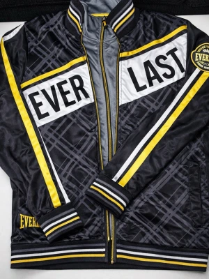 Set jacka och byxor/Everlast träningsset  - Stilrent och sportigt träningsset från klassiska Everlast, inspirerat av boxningens värld. Setet består av jacka och matchande byxor i svart med snygga detaljer i gult, vitt och grått. Jackan har hel dragkedja, ribbade muddar och krage samt tydlig EVERLAST-logotyp över bröstet. Byxorna har bekväm resår i midjan, dragkedjeförsedda fickor och ett broderat New York Everlast Boxing Dept-emblem som ger en exklusiv känsla. Passar perfekt för vardag och träning.  Storlek XS men passar även som S och M.