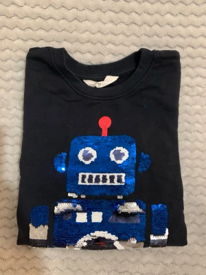 Svart tröja med paljettrobot H&M - Cool svart långärmad tröja från H&M med en stor blå robot framtill i glittriga paljetter. Rund halsringning och mjukt material som känns skönt mot huden. Perfekt för dig som gillar färgglada detaljer och vill sticka ut lite extra.