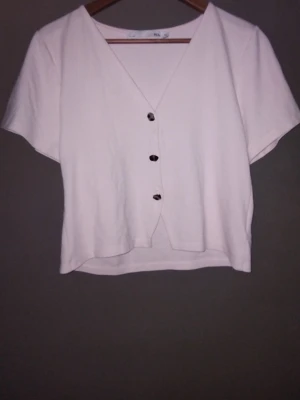 Ljusrosa blus med knappar från 157 - Kortärmad ljusrosa blus från 157 i storlek M. Toppen har v-ringning och fyra dekorativa knappar framtill. Tillverkad i mjukt bomullstyg och har en avslappnad passform som känns både enkel och stilren.