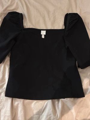 Svart topp med puffärmar från H&M - Säljer en svart topp från H&M med trendiga puffärmar och rak skärning. Toppen har en snygg, enkel design och är tillverkad i ett mjukt, stretchigt material som sitter bekvämt. Perfekt att styla med jeans eller kjol för en cool look.