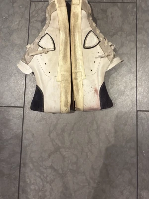 Vita sneakers från Philippe Model - Säljer ett par vita sneakers från Philippe Model med beige och svarta detaljer. Skorna har snörning, rund tå och platt sula. Ovandelen är i skinn och mocka, med den klassiska sköldloggan på sidan. Perfekta för dig som gillar en clean men ändå sportig look.