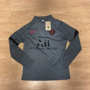 Grå PSG långärmad träningströja Nike - Snygg grå långärmad fotbollströja från PSG med svarta och röda detaljer. Tröjan har halv dragkedja, svarta mönstrade ärmar och tryck med 'Accor Live Limitless' och 'Visit Rwanda'. Perfekt för träning eller match. Tillverkad i funktionsmaterial. 