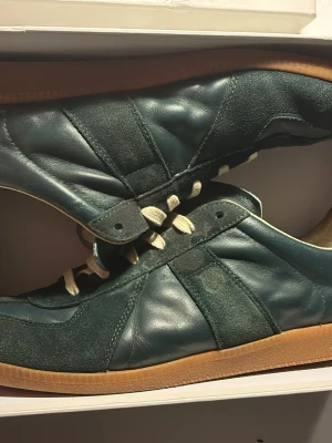 Mörkgröna maison margiela - Säljer ett par mörkgröna sneakers från maison margiela som har ett litet hål på vänster skon men det är inget stort hål och det kommer inte in något för här lagt en ny innersula bakom den vanliga. kommer med box har tyvärr inte kvitto 