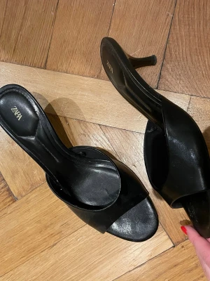 Svarta klackskor från Zara, storlek 40 - Säljer ett par stilrena svarta klackskor från Zara i storlek 40. Skorna har öppen tå, smal klack och bred rem över foten. Tillverkade i ett glansigt syntetmaterial som ger en elegant look. Perfekta för dig som gillar minimalistisk och trendig stil.