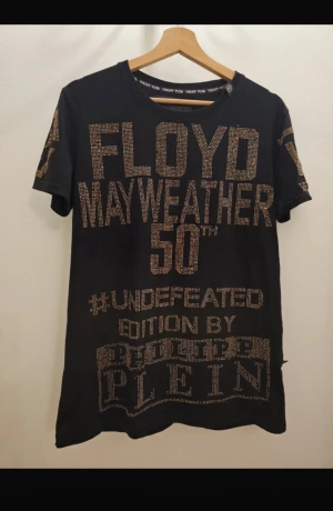 Philipp Plein x Mayweather t-shirt svart/guld - Svart t-shirt från Philipp Plein med stora guldiga stenar och texten 'Floyd Mayweather 50th #Undefeated Edition'. Kortärmad modell med rund hals och exklusiv design, perfekt för dig som vill sticka ut. Materialet är mjuk bomull och trycket är detaljerat både fram och bak. 