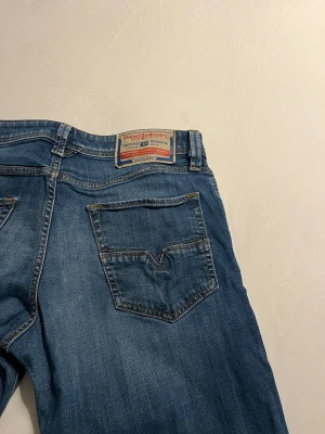 Blå raka jeans från Diesel - Snygga blå jeans från Diesel med klassisk femficksdesign och raka ben. Jeansen har en ljus tvätt med diskreta slitningar och detaljerade sömmar. Märkespatch bak i midjan och knappgylf framtill. Perfekta för en avslappnad och cool stil.