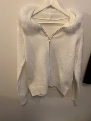 Vit jacka med fluffig huva - Säljer en vit jacka med dragkedja och fluffig pälsdetalj runt huvan. Tröjan har långa ärmar och är gjord i ett mjukt stickat material som känns riktigt cozy. Perfekt för dig som gillar en clean look med lite extra detalj på huvan.