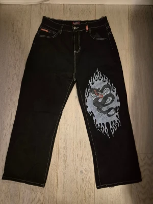Svarta Jnco jeans - Svarta streetware jeans med kontrastsömmar och en flames- och ormmålning på ena benet.