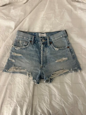 Ljusa denimshorts från Zara - Säljer ett par ljusblå denimshorts från Zara. Shortsen har slitna detaljer och klassisk femficksdesign. Perfekt för dig som gillar en avslappnad och trendig look.