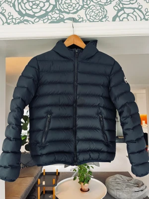 Svart Colmar vinterjacka med huva, stl 154 - Colmar boys Down Jacket, svart, storlek 154cm, 12 år, dunjacka med huva och två snedställda fickor med dragkedja. Jackan har ett stilrent, quiltat mönster och logotyp på ärmen. Perfekt för dig som vill ha en clean och varm look under kalla dagar. Vadderingen består av 90% dun och 10% fjäder. Normal passform.