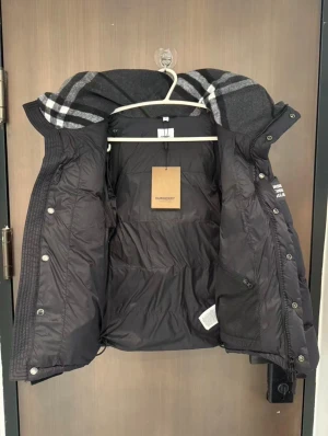 Svart pufferjacka från Burberry - Helt ny, aldrig använd, Svart pufferjacka från Burberry med klassisk rutig huva i grått och vitt. Jackan har dragkedja, tryckknappar och meshficka med dragkedja på insidan. Oversized look och quiltad design, perfekt för kalla dagar.