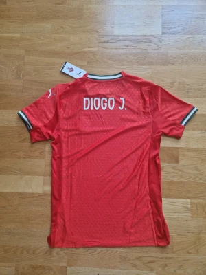 Portugal Diogo J. matchtröja Puma röd - Portugal fotbollströja med Diogo J. på ryggen, officiell Puma-modell. Röd med vita detaljer, broderad landslagssköld och Puma-logga. Kortärmad, lätt och ventilerande material med subtilt mönster och ribbade muddar i vitt och grönt.