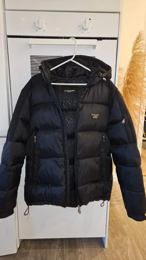Svart pufferjacka från Alessandro Zavetti - Svart pufferjacka från Alessandro Zavetti med huva och snygga detaljer som broderad logga på luvan och bröstet. Jackan har dragkedjor på fickorna och snörning nedtill. Perfekt för kalla dagar med sin tjocka och quiltade design.
