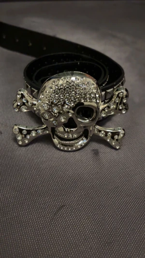 Svart nitarbälte med dödskalle-spänne - Coolt svart bälte i skinn med silverfärgade nitar längs hela bandet och ett stort dödskalle-spänne dekorerat med glittrande stenar. Perfekt accessoar för dig som vill sticka ut och addera lite edge till din outfit.
