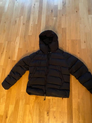 Svart pufferjacka från C.P. Company - Säljer en svart pufferjacka från C.P. Company med ikoniska goggles i huvan. Jackan har en schysst quiltad design, dragkedja framtill och två stora fickor. Perfekt för dig som vill ha en snygg och funktionell vinterjacka med streetkänsla.