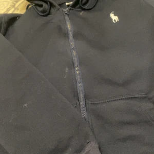 Mörkblå hoodie från Polo Ralph Lauren - Snygg mörkblå hoodie från Polo Ralph Lauren med klassisk vit logga på bröstet. Tröjan har huva, dragkedja framtill och praktiska fickor. Perfekt för chill dagar och enkel att matcha med jeans eller joggers.