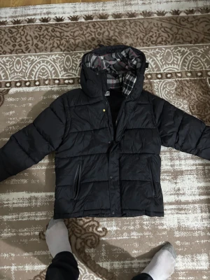 Svart pufferjacka från Burberry - Säljer en svart pufferjacka från Burberry med stor huva och tydlig logga på ärmen. Jackan har dragkedja, knappar och två fickor framtill. Insidan av huvan har rutigt foder i grått, rött och vitt. Perfekt för kalla dagar och riktigt snygg streetstil. Byte funkar